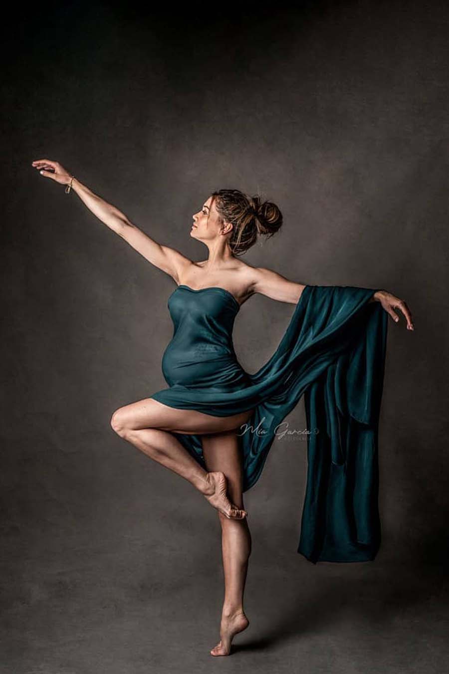 This pregnant model is doing a ballerina pose. She has her arms wide and is standing on one leg. She has the fabric of the silky scarf around her upper body. The rest of the fabric is hanging on her stretched arm.