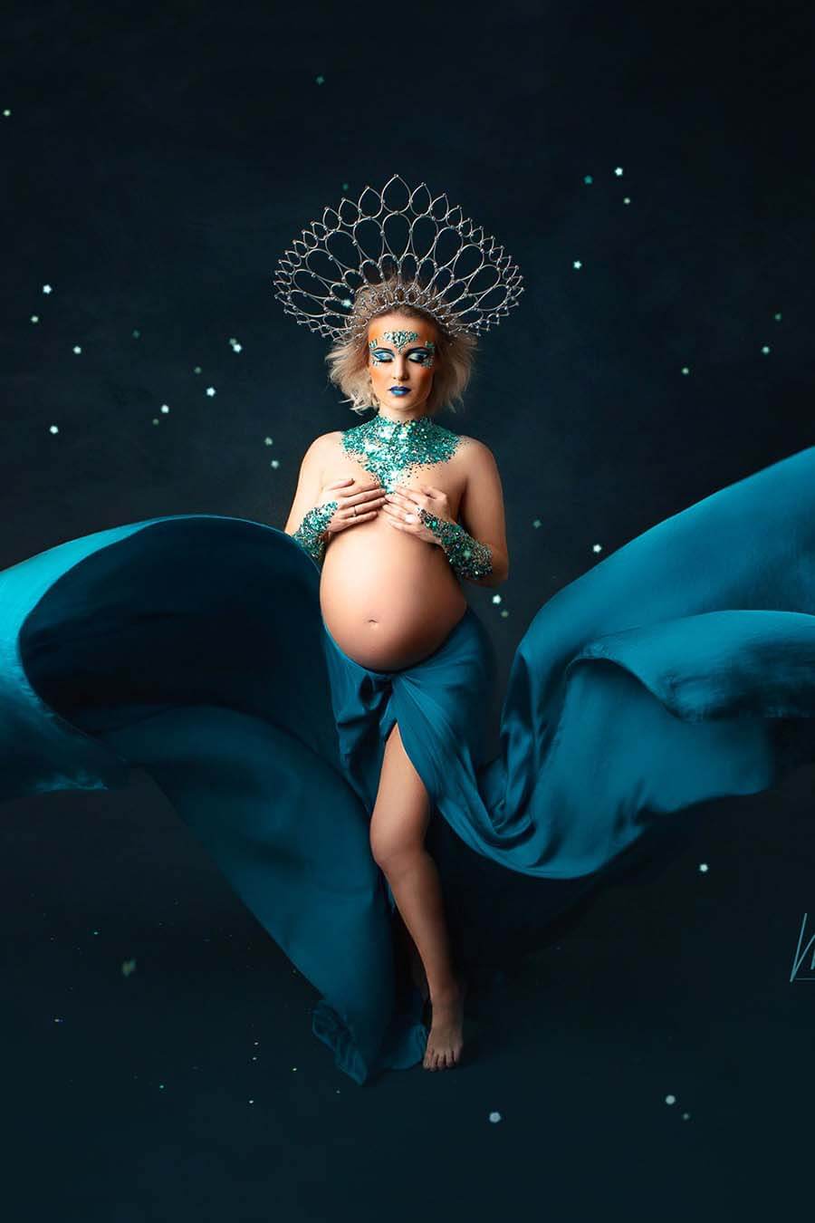 A pregnant woman is posing for the photo. They tied the silky scarf petrol to a skirt with a split by the leg. She is covered in glitters and little diamant stones on her underarms, neckline and her face. She is wearing a huge crown in her hair.