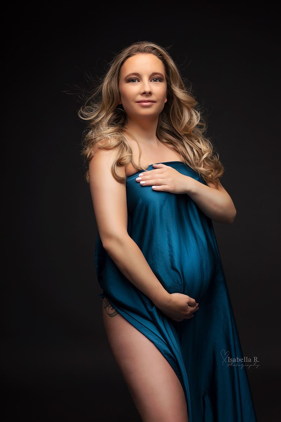 A pregnant model with blonde hair is in a studio. She is holding up the silky scarf in front of her. She has one hand underneath her belly. 