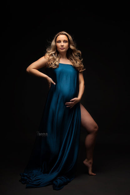 A pregnant model is posing for the photograph. She is wearing a silky scarf. She has one leg out of the fabric. The model has blond hair with waves. She is posing with one hand in her side and the other one is underneath her belly.