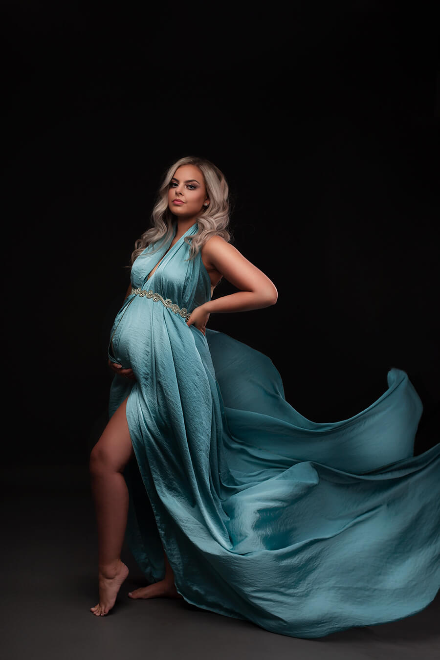 Blond pregnant model wears the mii-estilo 4 meter long silky scarf in powder blue color. The scarf is dressed on the model and adorned with a sash as accessory. The model faces the camera  with a side look. One hand holds her bump and the other holds her back. 