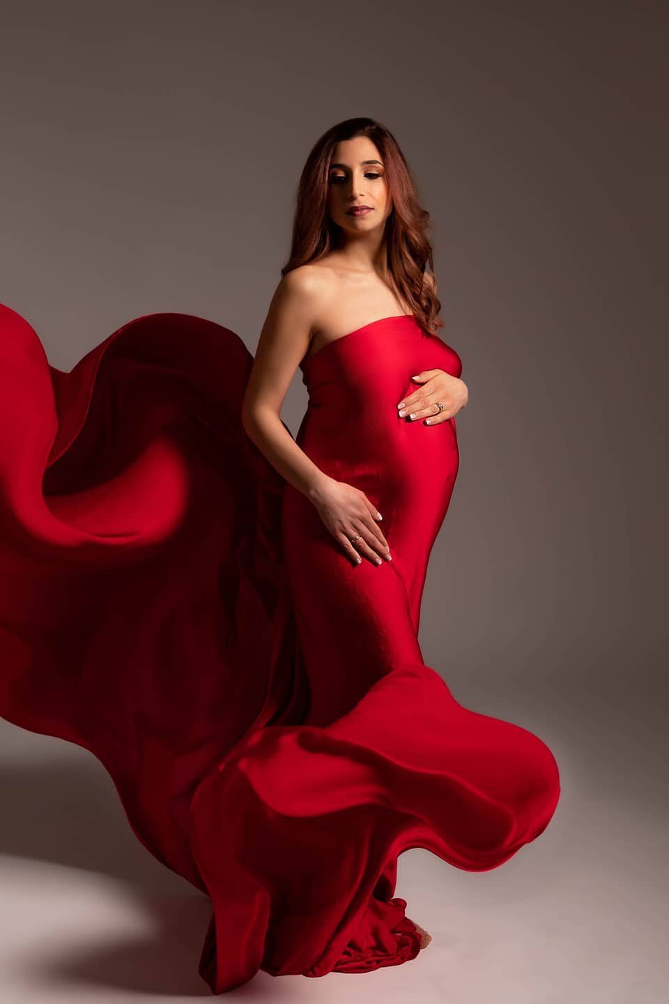 Red haired pregnant model poses with a scarf covering her body. The scarf is very long, made of silk in a vibrant red color.