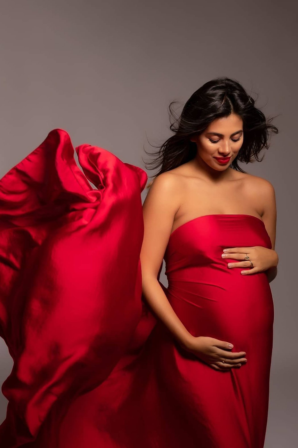Brunette model poses with her eyes closed touching her bump. She is wearing a vibrant red long silky fabric. 