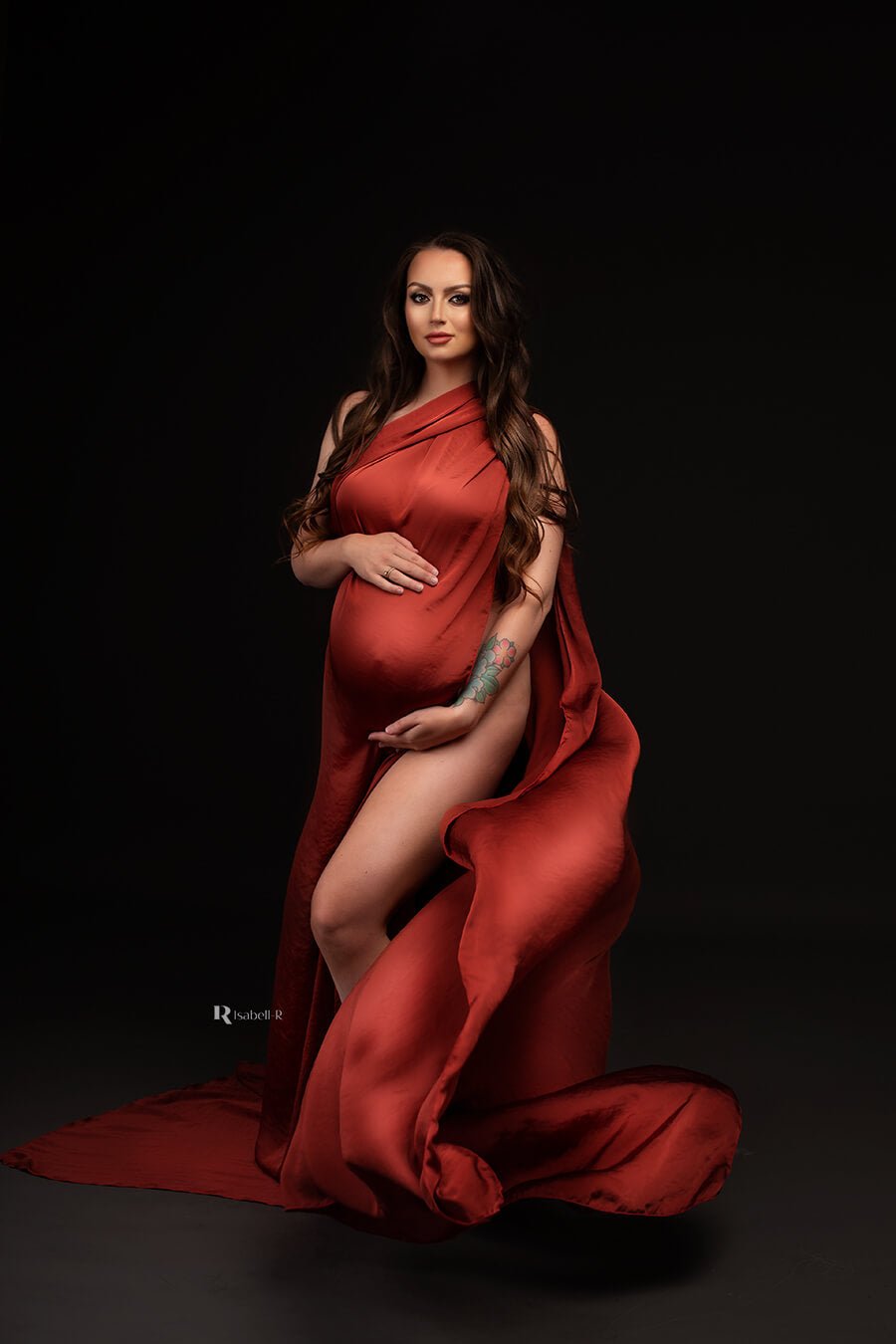 Brunette model poses in a studio during a maternity photoshoot. She wears only a scarf to cover her body. She holds her bump with both of her hands. The model faces the camera and the scarf is thrown just before the shot, the photo captures the moment that the rust orange scarf falls in a playful way.