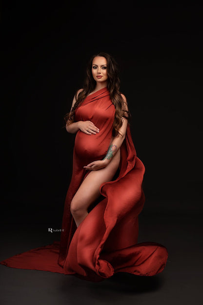 Brunette model poses in a studio during a maternity photoshoot. She wears only a scarf to cover her body. She holds her bump with both of her hands. The model faces the camera and the scarf is thrown just before the shot, the photo captures the moment that the rust orange scarf falls in a playful way.
