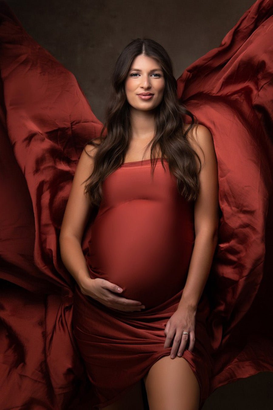 brunette pregnant model poses in a studio with a silky scarf in rust orange wrapped around her body.