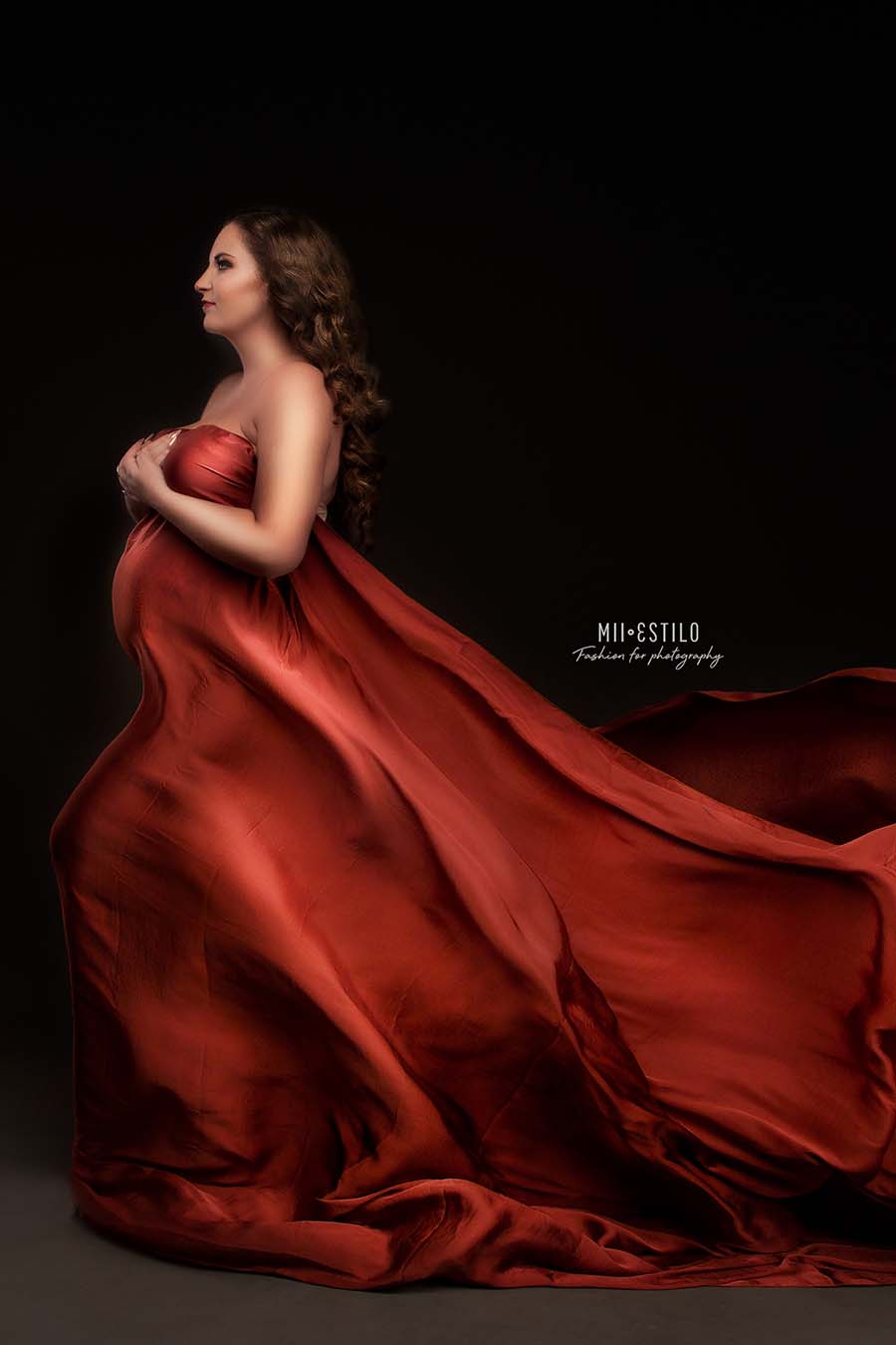 pregnant model poses in a studio against a dark background wearing a rust orange silky scarf to cover her body. she is posing on her side and holds the scarf against her body.