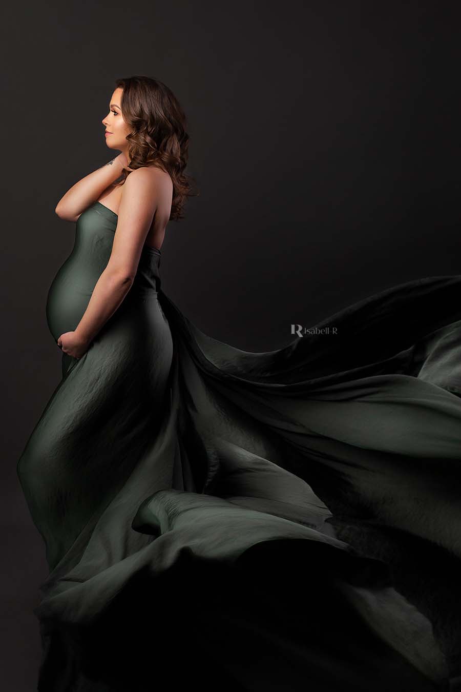 model poses on her side with her eyes open touching her neck and holding her bump with the other hand. she is wearing a long silky draping fabric that is covering her body,