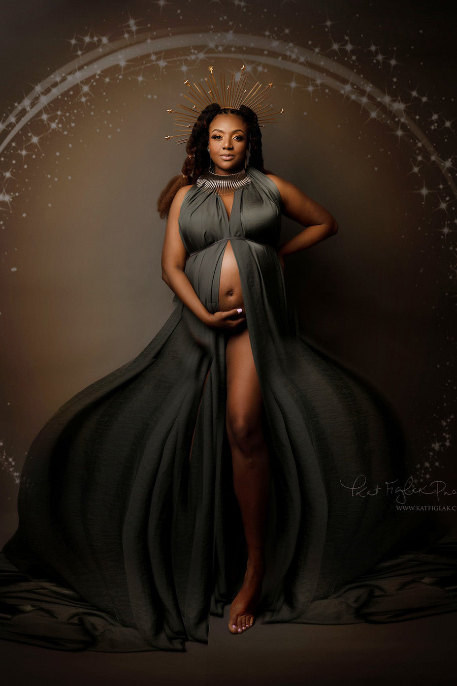pregnant model poses against a brown background with stars. she is wearing a slategrey silky scarf wrapped around her body as a dress. she has a crown and a big necklace to match the style. 