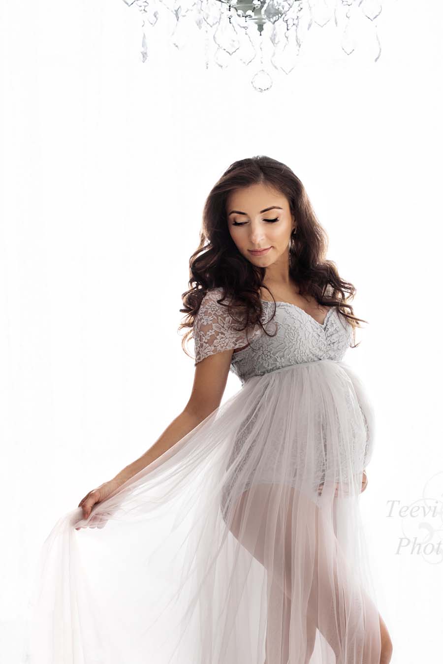 pregnant model poses with her eyes closed in a white studio holding a tulle train that is attached to her lace bodysuit in cool grey color.