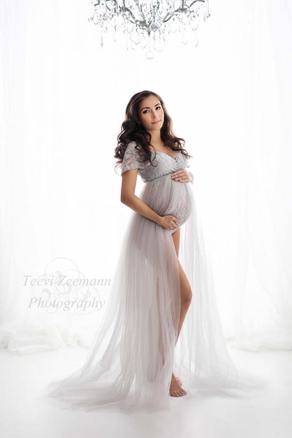 model poses in a white studio wearing a cool grey set made of a lace bodysuit and a tulle skirt. she is staring at the camera and holding her bump with both hands.