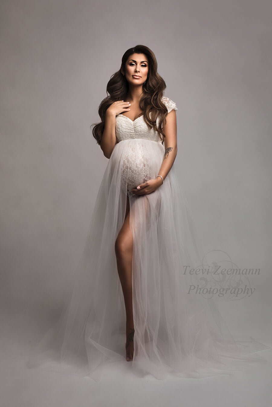 Brunette model poses in a studio wearing the strelitzia set from mii-estilo. The off white gown is a bodysuit together with a tulle skirt. The model is standing tall and facing the camera - one hand holds her bump and the other lays on her neck. One of her legs can be seen through the tulle skirt split.