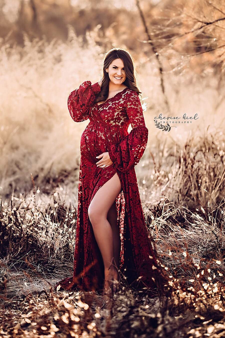 A pregnant woman is standing outside in the nature. She is wearing a bordeaux red lace dress. She is smiling. the dress has a split by her leg