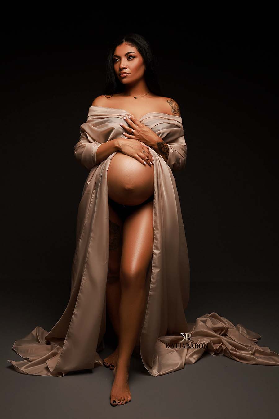 pregnant model poses in a dark studio wearing a sand taffeta scarf covering her chest and leaving her belly uncovered.