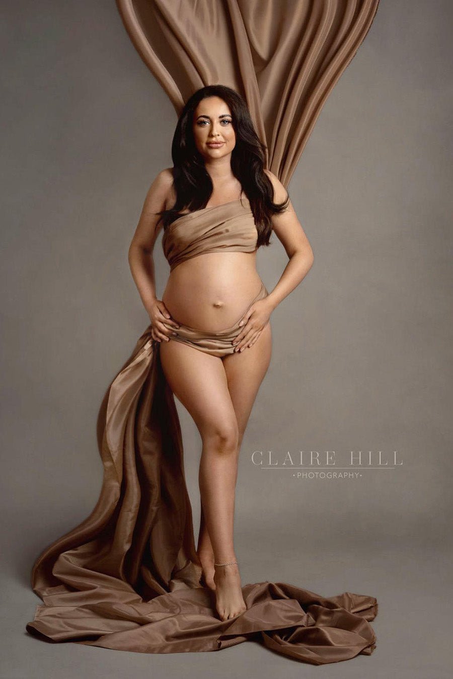 pregnant model with long dark hair poses in a studio with a taffeta scarf in sand color wrapped around her body. 