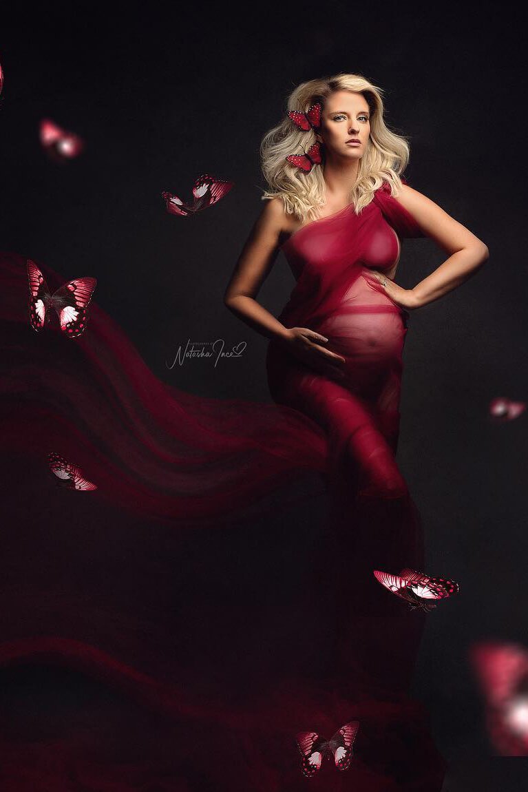 A pregnant model is using a tulle scarf as a dress. The model has short blond hair. The fabric is tied around her shoulder to create a one shoulder dress effect. The fabric is tight around her and lose next to her. She has two butterflies in her hair and there are more of them flying around.