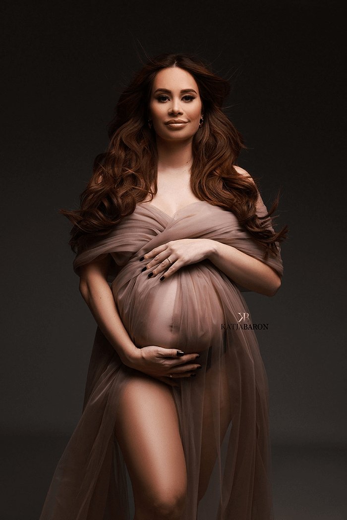 Pregnant brunette model wears a draping tulle fabric in sand color wrapped on her shoulders and covering her belly. She holds her bump with both hands and stares at the camera. 