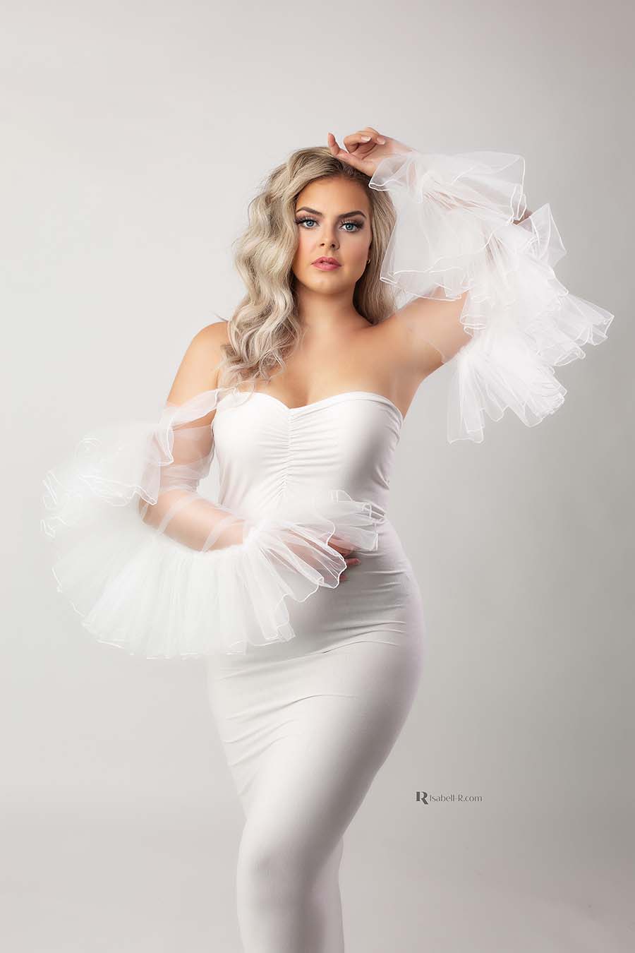 Blond model poses in a studio wearing a tight white dress and matching tulle sleeves. one hand lays on her head and the other on her belly. 