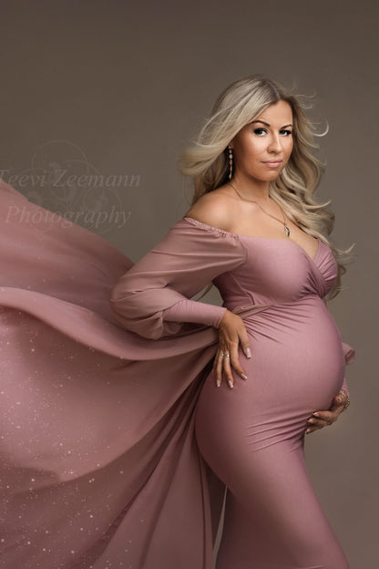 Blond pregnant model poses in a studio during a maternity photoshoot. She looks down with her eyes partially closed. She wears an old pink long dress designed by mii-estilo. The dress features a tight underdress with long chiffon train. The top has a sweetheart neckline and long bell sleeves. The model holds her bump with one hand and the other is laying on her waist. The train is being handheld by someone else to capture flying effects during the shot. 