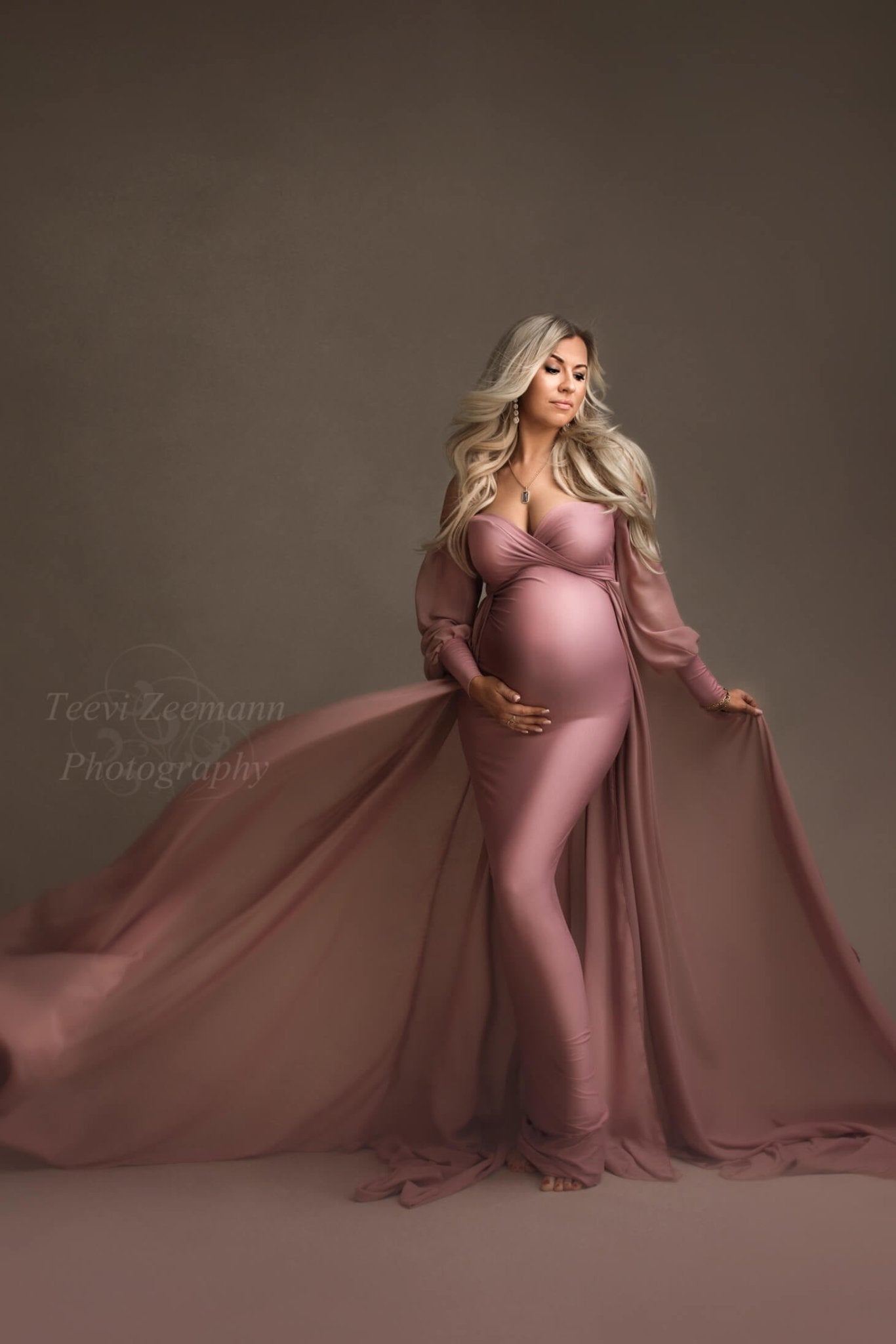 Blond pregnant model poses in a studio during a maternity photoshoot. She looks down with her eyes partially closed. She wears an old pink long dress designed by mii-estilo. The dress features a tight underdress with long chiffon train. The top has a sweetheart neckline and long bell sleeves. The model holds her bump with one hand while playing with the train with the other hand.