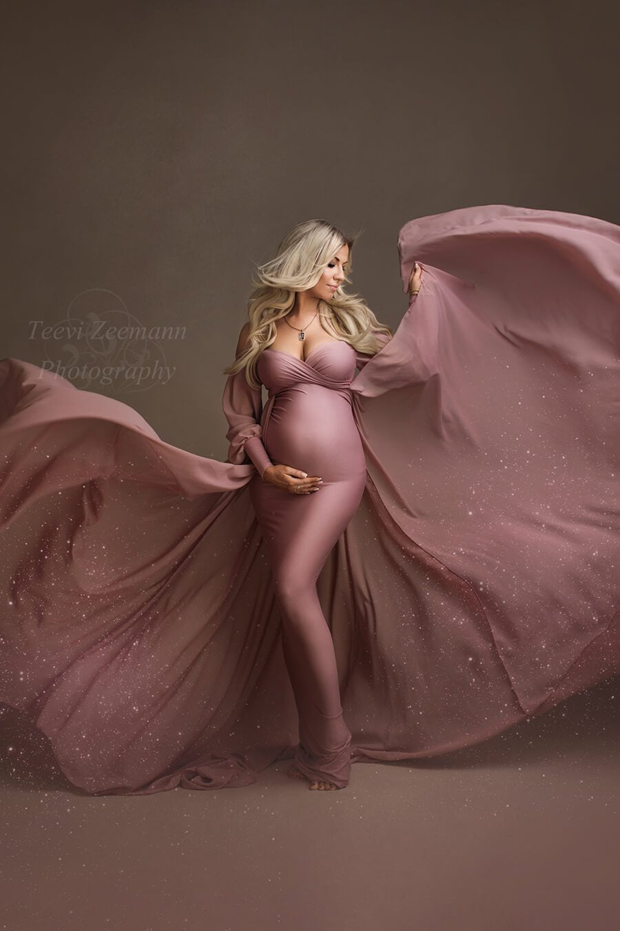 Blond pregnant model poses in a studio during a maternity photoshoot. She looks down with her eyes partially closed. She wears an old pink long dress designed by mii-estilo. The dress features a tight underdress with long chiffon train. The top has a sweetheart neckline and long bell sleeves. The model holds her bump with one hand while playing with the train with the other hand. 