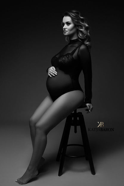 A pregnant model is sitting on a stool. She is facing to the side. She has long nails.The bodysuit she is wearing is black and made from mesh. She has curls in her hair