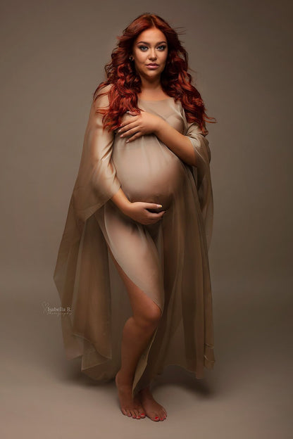 A pregnant woman with bright red hair is wearing a espresso cape. She is looking towards the camera and has her hand on her belly