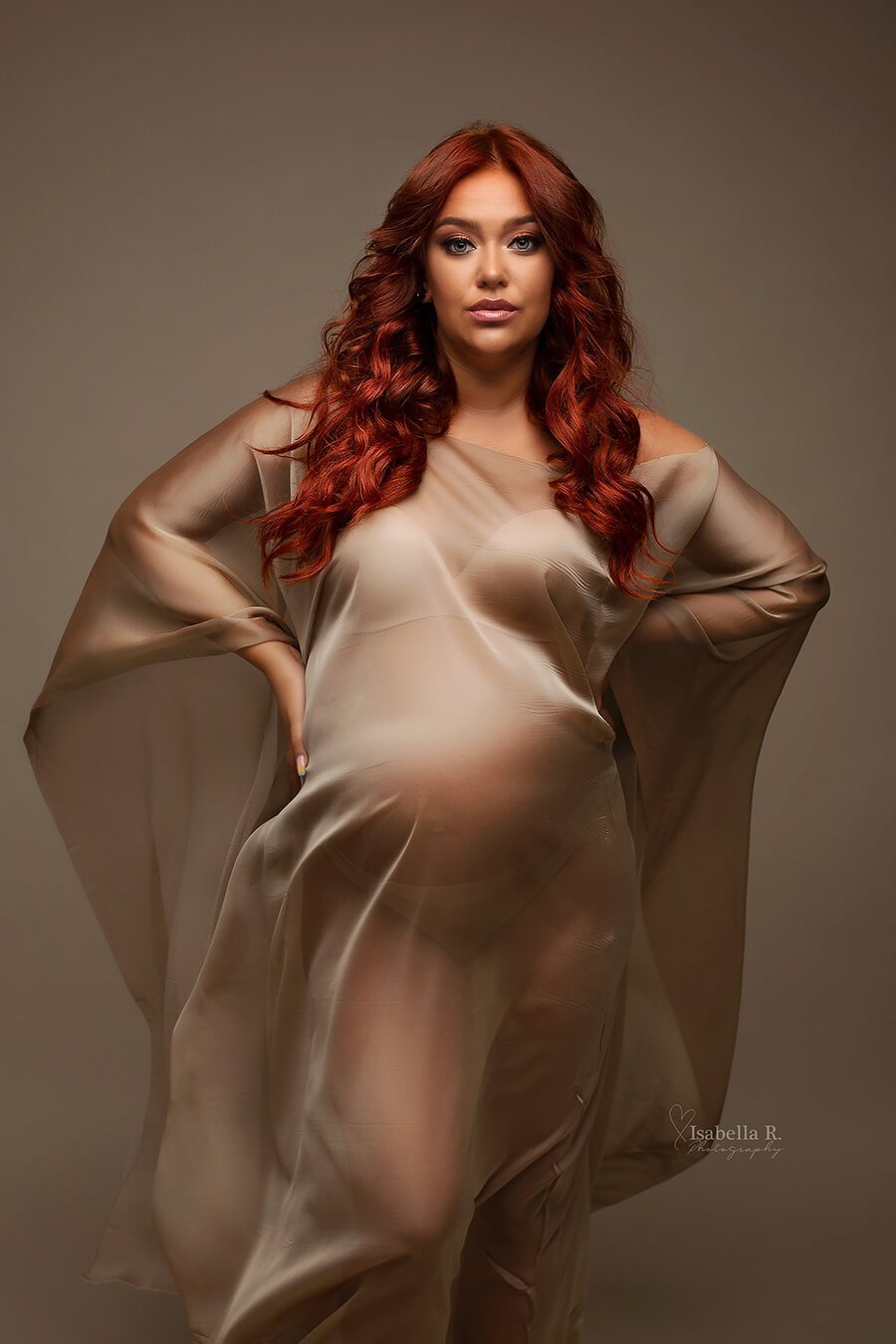 A pregnant woman with red hair is wearing a espresso cape. the cape is hugging her body because of the wind