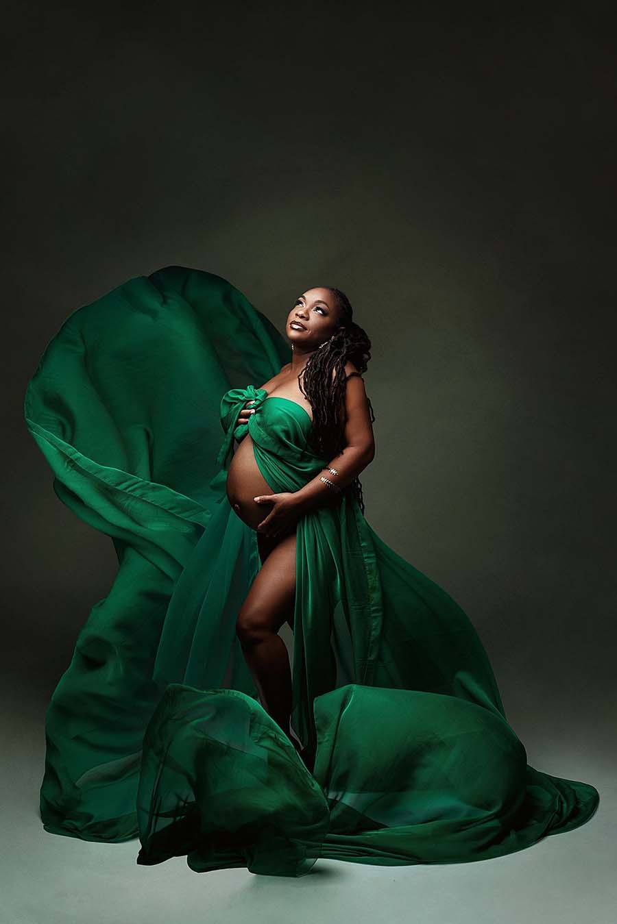 Pregnant model poses in a studio wearing a draping fabric in green color covering her chest area and leaving belly and legs uncovered. She faces up while holding the scarf against her chest with one hand and her bump with the other. 