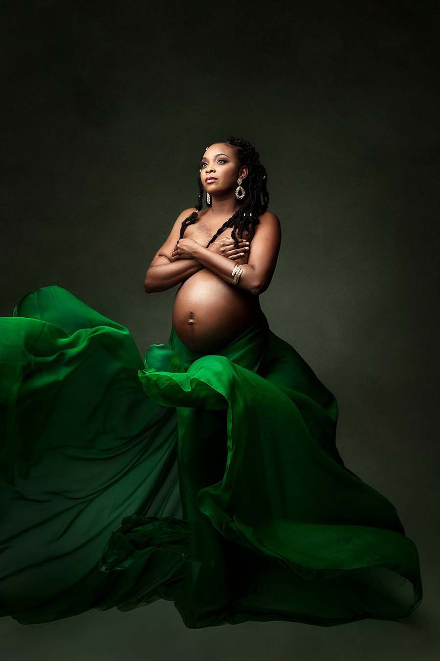 Pregnant model poses in a studio wearing a green scarf tied in her waist. she has her hands covering her breasts and is looking up. 