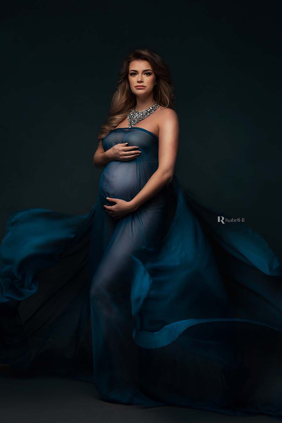 A pregnant model is wearing a voile scarf as a dress. The color of the scarf is petrol and the fabric is a little bit sheer. She is wearing a big statement necklace to finish the look. 