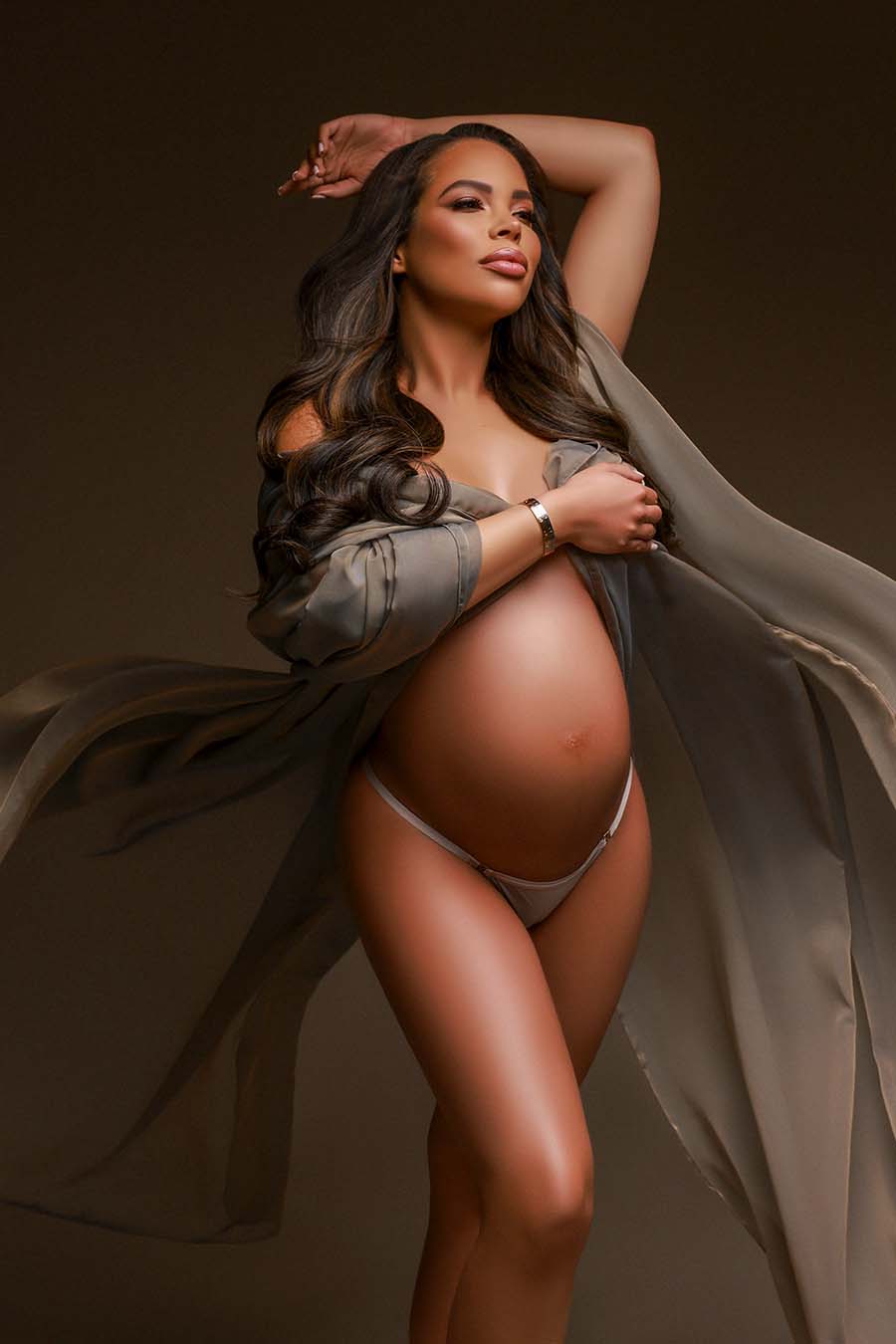 Pregnant model poses during her maternity photoshoot wearing a draping fabric made of voile in taupe color. she cover her breasts with the scarf and has her belly uncovered. She has her eyes closed and one hand above her head during the close up.