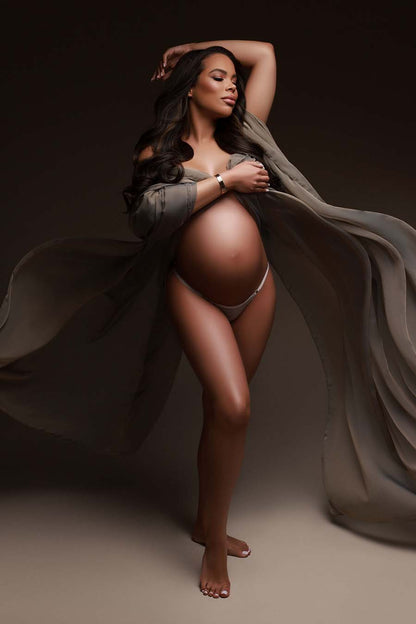 Pregnant model poses during her maternity photoshoot wearing a draping fabric made of voile in taupe color. she cover her breasts with the scarf and has her belly uncovered. She has her eyes closed and one hand above her head during the close up. 