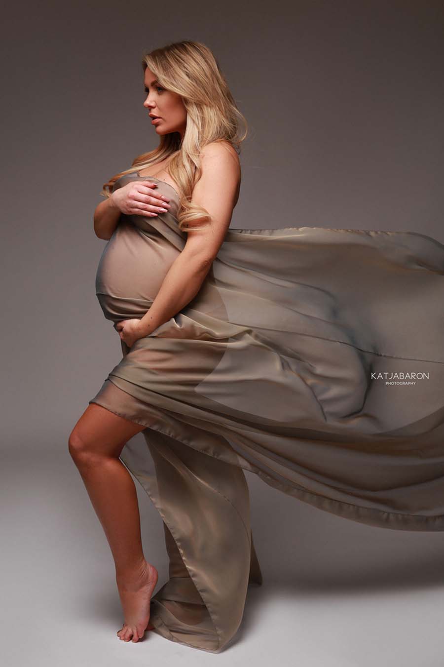 A blond model that is pregnant has a scarf around her. The extra fabric of the scarf is used to create a wave behind her. 