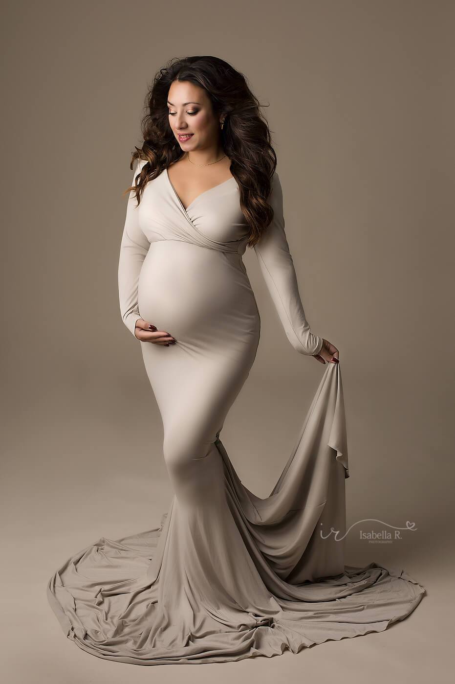 A pregnant woman is wearing a tight dress in the colour sand. The dress has a mermaid skirt. The woman is holding a piece of the skirt fabric in her hand. She has brown hair
