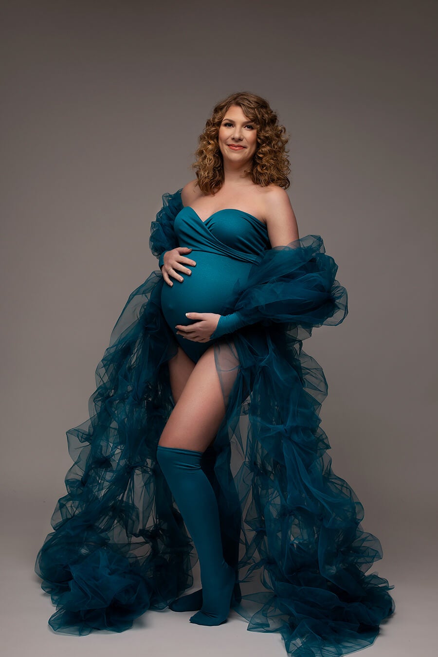 A pregnant woman is posing in a studio She is wearing The woman is wearing high socks, loose sleeves and a bodysuit with a bandeau top. Everything is in the color petrol. She has a tulle scarf by her arms that is going around her back. The rest of the tulle is besides her till the groud.
