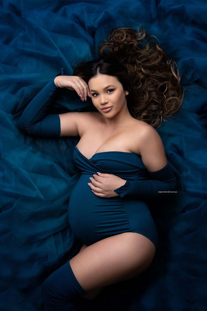 A pregnant model is laying on the ground. She is looking upwards to the camera above her. She is laying on tulle fabric. She is wearing a 3 piece set. There are two sleeves with a point at the end. The bodysuit has a bandeau top. The finish the look she is wearing two high socks. The color of the set is Petrol.