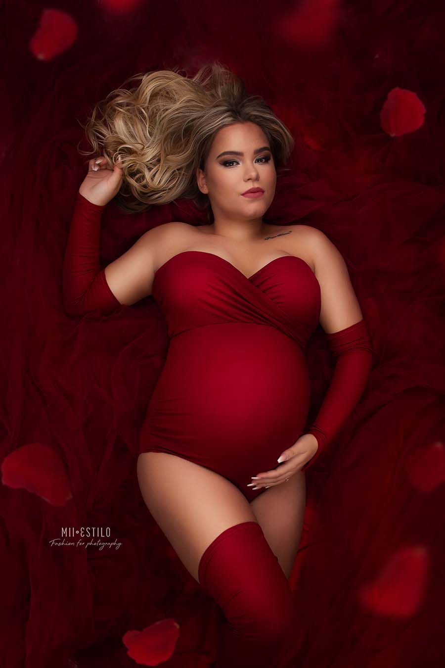 blond pregnant model poses laying on the studio floor wearing a maternity set consisting in a bodysuit, strapless with an adjustable sweetheart top, gloves and socks - all in red color. the model is laying on a big tulle piece, in red color as well.